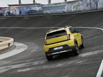 Een gele Fiat Grande Panda SUV met kenteken "FI 000 AI" rijdt op een gebogen, hellende weg met een muurschildering op de achtergrond.