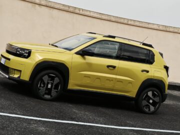 Een gele Fiat Grande Panda SUV staat geparkeerd op een zwart asfaltoppervlak, grenzend aan een hoge beige muur.