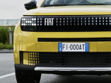 Vooraanzicht van een gele Fiat Grande Panda met een Europees kenteken "FI-000AT", geparkeerd op een verhard terrein naast een lage heg.