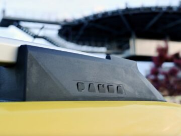 Close-up van een zwart-gele Panda-imperiaal op een Fiat Grande Panda. Op de achtergrond is een wazig, groot bouwwerk van metalen balken en platforms zichtbaar.