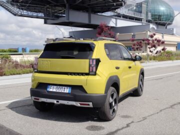 Een gele Fiat Grande Panda SUV staat geparkeerd vlakbij een modern gebouw met een bolvormige glasstructuur.