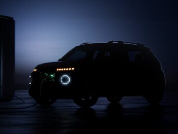 Die Silhouette eines Hyundai INSTER Elektro-Geländewagens wird an einer Station unter dem Nachthimmel aufgeladen, wobei die Scheinwerfer abgeblendet sind. Die Ladestation verfügt über eine leuchtende grüne Kontrollleuchte, die eine stimmungsvolle Szene erzeugt.