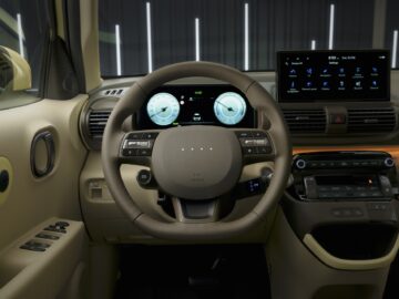 Ein Hyundai-Autoarmaturenbrett mit einem Lenkrad, einer digitalen Instrumententafel und einem Infotainment-Bildschirm mit mehreren Bedienelementen. Das Interieur des Hyundai INSTER ist in Beige- und Schwarztönen gehalten und verfügt über zahlreiche Tasten und Lüftungsöffnungen.