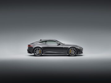 Side view of a sleek, silver Jaguar F-Type sports car with aerodynamic design, yellow brake calipers and stylish alloy wheels, set against a plain, gradient background.