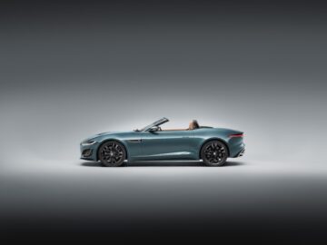 A sleek, blue Jaguar F-Type convertible sports car with the top down, seen from the side, against a simple gray background.