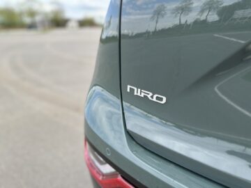 Close-up van de achterkant van een groen voertuig met het embleem "Niro" zichtbaar, wat aangeeft dat het een Kia Niro Electric is. De achtergrond is een buitenruimte met een verhard oppervlak en vage bomen.