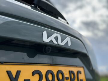 Close-up van de achterkant van een grijze Kia Niro Electric-auto, met het Kia-logo en een deel van de kentekenplaat met registratie "X-209-PG." De lucht is bewolkt op de achtergrond.