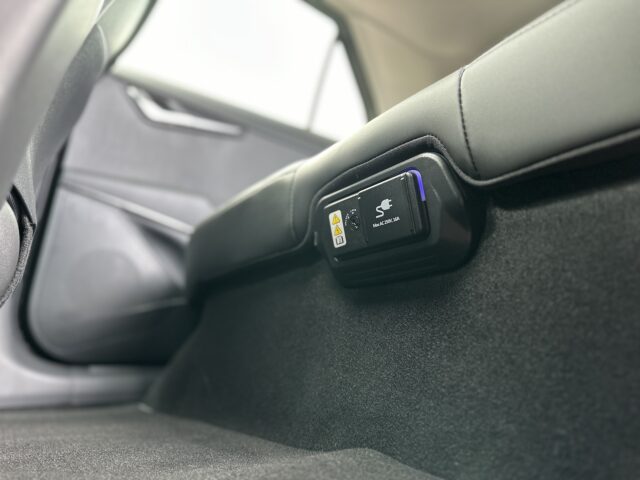Een close-up van de achterbank van een Kia Niro Electric, gefocust op een zwart elektronisch apparaat dat onder de stoel is gemonteerd met het label 'Seat Sensor Air'. De autodeur en vloer zijn gedeeltelijk zichtbaar.