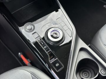 Close-up van de middenconsole van een Kia Niro Electric met een draaiknop, verschillende bedieningsknoppen, een bekerhouder en een deel van de autostoel.