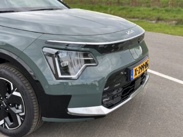 Close-up van de voorkant van een groene Kia Niro Electric met Nederlands kenteken, met opvallende koplampen en een Kia-logo op de motorkap. De auto staat geparkeerd op een verhard terrein met groen gras op de achtergrond.