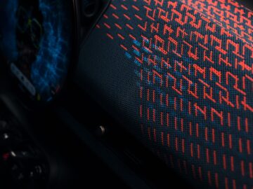 Close-up van het dashboard van de Aceman met een rood en blauw structuurpatroon op een donker oppervlak, met een deel van een digitaal scherm en enkele bedieningsknoppen zichtbaar aan de linkerkant, wat het strakke ontwerp van deze elektrische MINI benadrukt.