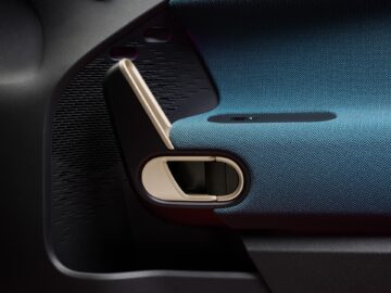 Close-up van een binnendeurpaneel van een auto met een blauw beklede armsteun, een strakke deurgreep en een kleine bedieningsknop, die de luxueuze details van het ontwerp van de Aceman laat zien.