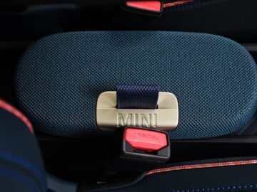 Close-up van een MINI-autogordelgesp, voorzien van een gouden plaat met het woord 'MINI' gegraveerd, geplaatst op een blauwgroene stoelbekleding met structuur en rode stiksels. Dit detail weerspiegelt het premium design van elk elektrische MINI-model.