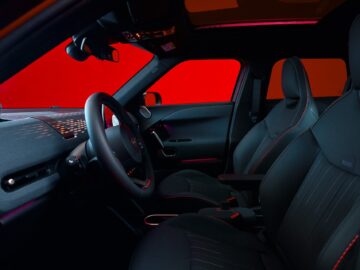 Interieur van een elektrische MINI Aceman met zwarte stoelen met rode stiksels, een zwart stuur en rode sfeerverlichting. Het dashboard toont een digitale interface. De achtergrond is rood verlicht.