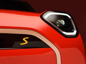 Close-up van de voorkant van een rode elektrische MINI, met een koplamp en een deel van de grille, met een geel "S"-embleem.