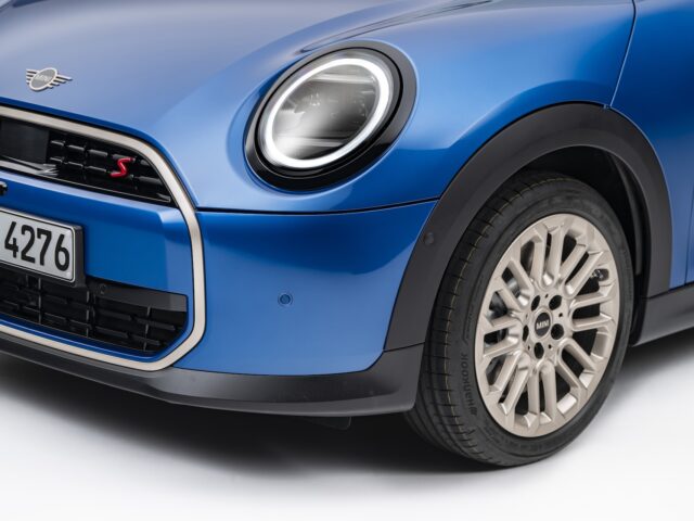 Close-up van het rechtervoorgedeelte van een nieuwe 5-deurs MINI Cooper, met de nadruk op de koplamp, de grille en het stuur met een kentekenplaat met de tekst "SEI 4276.