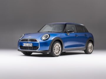 Een nieuwe blauwe MINI Cooper 5-deurs hatchback staat geparkeerd in een studio met een grijze achtergrond, waarbij de voor- en besturing van de auto worden getoond. De auto heeft een Duitse kentekenplaat met "M-EL 4276".