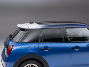 Zijaanzicht van een blauwe compacte auto met 5 deuren, zwart getinte ramen en een wit dak, tegen een effen grijze achtergrond, die het kenmerkende ontwerp van een MINI Cooper laat zien.