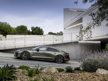 Een strakke, grijze Audi RS e-tron GT geparkeerd op een moderne betonnen oprit, omgeven door groen en architectonische planten, met een modern huis op de achtergrond, dat zijn prestatiepotentieel van 1.000 pk laat zien.