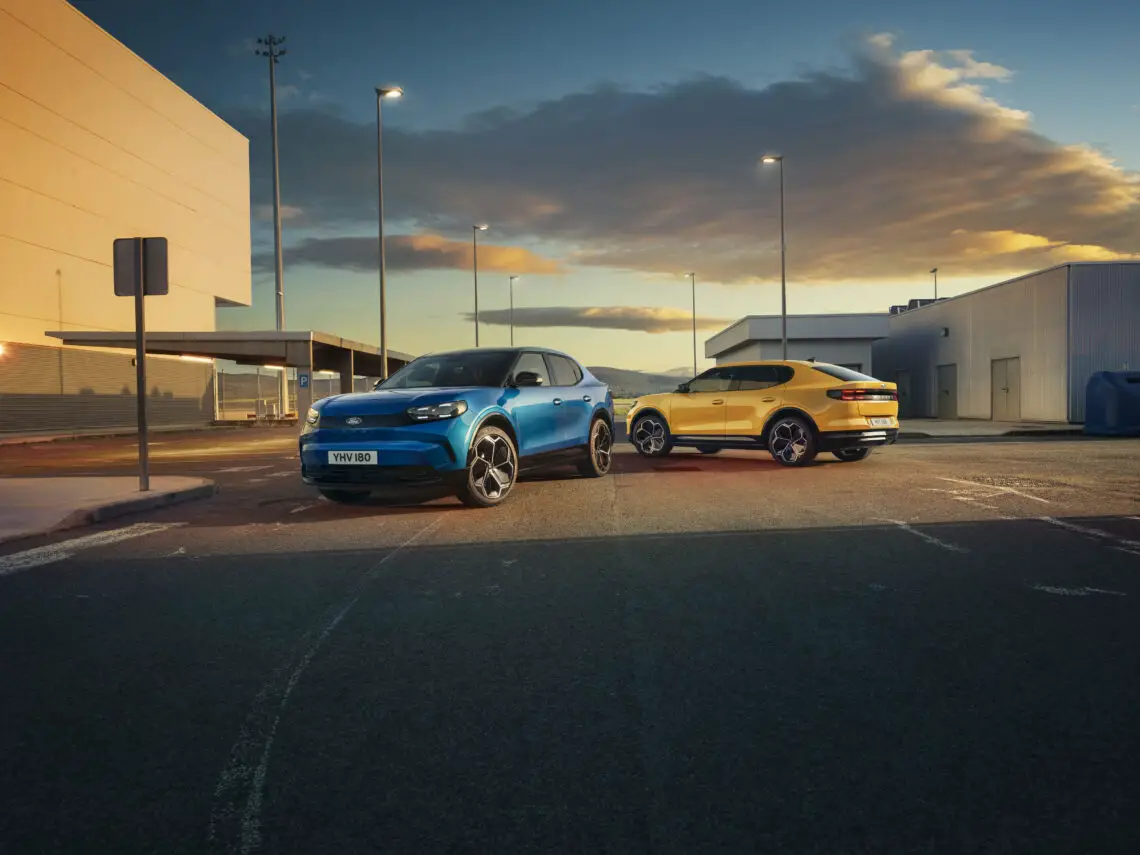 Twee moderne auto's, een blauwe en een gele, geparkeerd op een industriegebied onder straatverlichting bij zonsondergang, die doet denken aan de klassieke Ford Capri in een tijdloze setting.