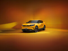 Een gele SUV, die doet denken aan de klassieke charme van een Ford Capri, wordt tentoongesteld in een studio met een oranje achtergrond. Op het kenteken van de auto staat 
