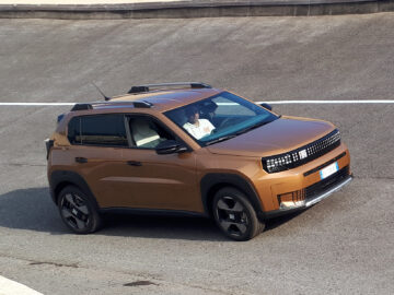 Un todoterreno Fiat Grande Panda de color bronce circula por una carretera asfaltada, con el interior reluciente a la luz del sol, mientras una persona con camisa blanca conduce con confianza.