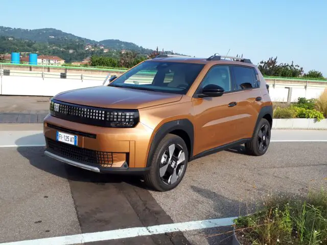 Ein brauner Fiat Grande Panda Geländewagen parkt auf einem gepflasterten Parkplatz mit einer grünen Landschaft und blauem Himmel im Hintergrund. Das Fahrzeug hat ein modernes Styling mit einem schwarzen Kühlergrill, einem markanten Raddesign und einem gut gestalteten Innenraum.