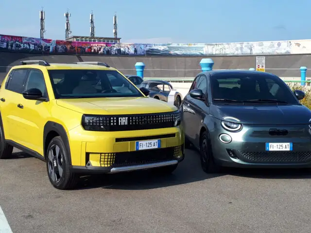 Op een asfaltoppervlak staan twee auto's naast elkaar geparkeerd, met links een gele SUV en rechts een grijze Fiat Grande Panda. De achtergrond toont een licht gebogen, verhoogde baan.