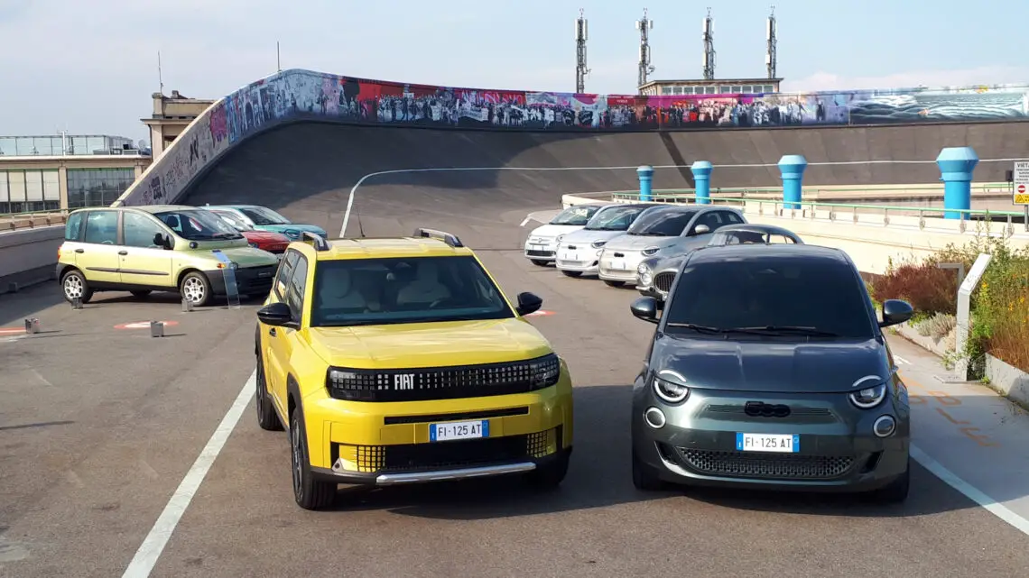 Deux voitures garées, un SUV jaune à gauche et une voiture compacte gris foncé à droite, se trouvent dans un parking sur le toit, avec plusieurs autres voitures et une piste en pente à l'arrière-plan. Parmi elles se trouve une Fiat Grande Panda, dont l'intérieur présente un design épuré tout en incorporant des groupes motopropulseurs efficaces.