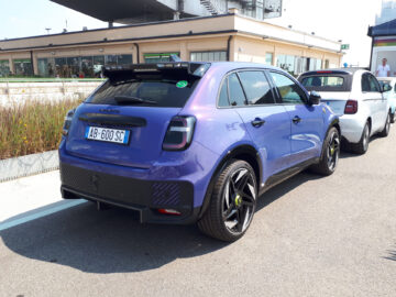 Een blauwe hatchback-auto staat aan de kant van een straat geparkeerd, met andere voertuigen zichtbaar op de achtergrond. De auto, die doet denken aan klassieke Fiat modellen, heeft een kenteken met de tekst "AB-600-SC".