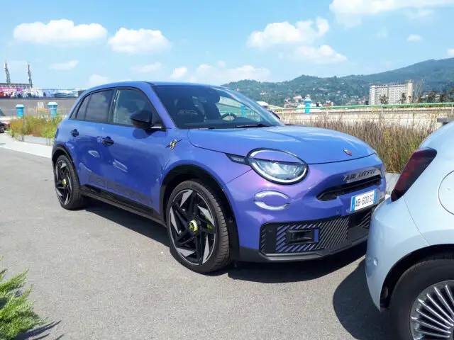 Een blauwe Abarth SUV, een knipoog naar de klassieke modellen van Fiat, staat naast een lichtblauw voertuig geparkeerd op een asfaltweg, met op de achtergrond uitzicht op heuvels en gebouwen onder een heldere hemel.