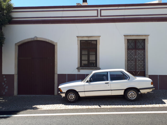 Een klassieke, vroege BMW 316 staat geparkeerd op een geplaveide straat voor een gebouw met witte muren, twee ramen en een gesloten garagedeur.