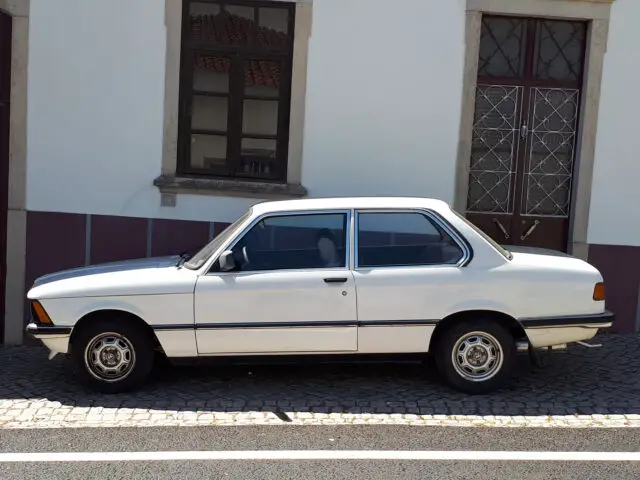 A white classic BMW 316 is parked on a cobblestone street in front of a building with two windows and a door. Spotted during an early morning drive, this timeless vehicle exudes elegance and charm.