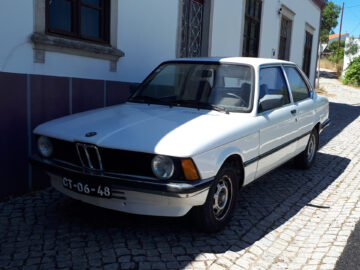 Une BMW 316 berline blanche d'époque est garée dans une rue pavée, à côté d'un bâtiment à deux fenêtres. Le numéro d'immatriculation de la voiture indique 