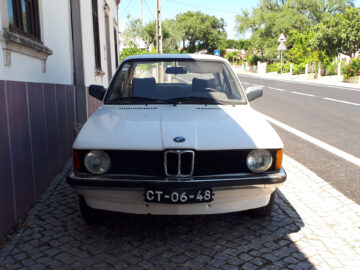 Un BMW 316 blanco y antiguo está aparcado en una calle asfaltada junto a un edificio de base morada, cerca de una zona residencial. En la matrícula se lee 