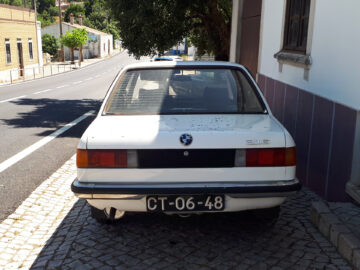 La parte trasera de un BMW 316 antiguo, un coche blanco con matrícula CT-06-48, está parcialmente aparcada en una acera pavimentada junto a un edificio y una carretera. Visto en un entorno urbano, este vehículo clásico llama la atención.