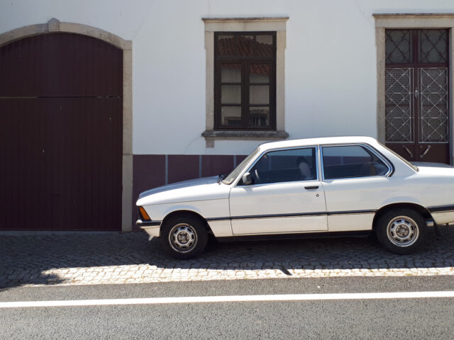 Een vroege BMW 316 staat geparkeerd in een geplaveide straat voor een wit gebouw met een gesloten houten garagedeur en twee ramen, die de essentie van de vintage charme van Gespot vastleggen.