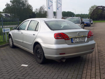 Een zilveren Brilliance BS4 sedan met een "NSZ 00300" kentekenplaat staat geparkeerd op een verhard terrein nabij een metalen hek en bewegwijzering. Bomen en andere voertuigen zijn zichtbaar op de achtergrond, wat erop wijst dat het recentelijk is gespot.