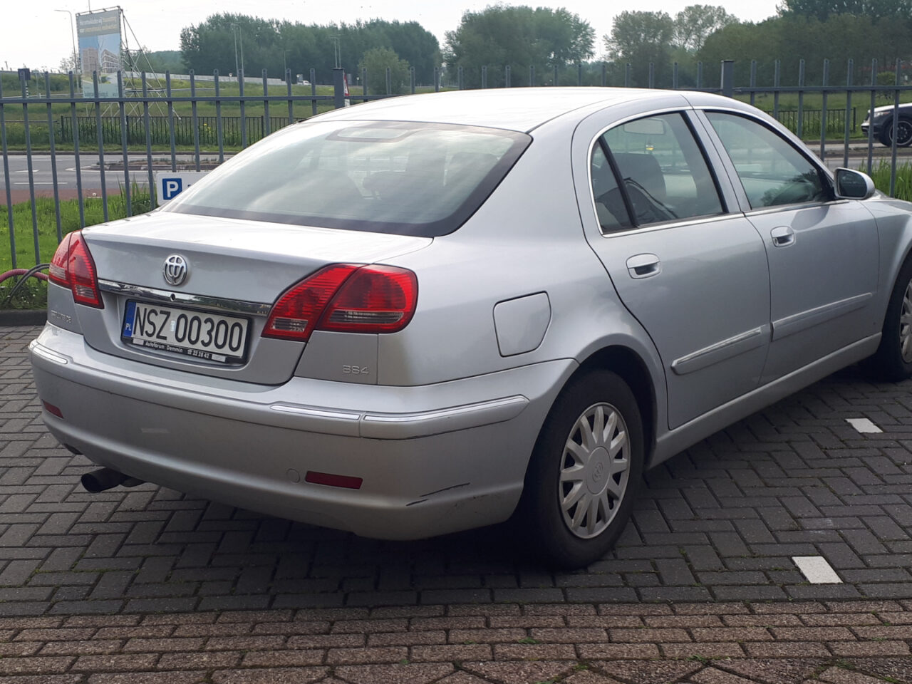 Een zilveren Brilliance BS4 sedan geparkeerd op een parkeerplaats met een Europees kenteken "WSZ 00300", naast een zwarte auto en een metalen hek. Op de achtergrond zijn bomen en een weg zichtbaar.