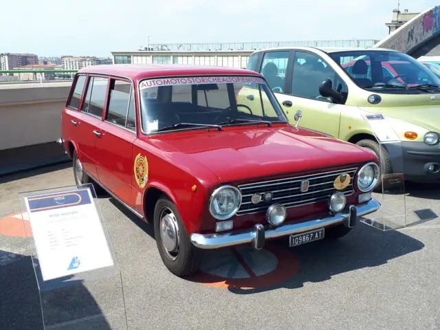 Buiten staat een rode vintage stationwagen, naast een groene auto. Op de voorruit staat de tekst "AUTOMOTO STORICHE PIEMONTE", en een bord geeft details over het model en de geschiedenis van de auto. Deze klassieke Fiat voegt charme toe aan de tentoonstelling met zijn tijdloze design.