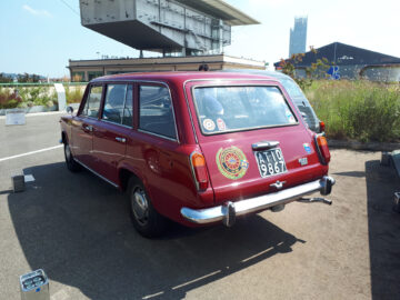 Een klassieke rode Fiat 124 stationwagen, een van de tijdloze klassieke Fiat-modellen, staat buiten geparkeerd bij een modern gebouw. De achterkant van de auto is voorzien van diverse stickers en een kentekenplaat van de Europese Unie, waardoor de charme van klassieke auto's in een eigentijdse setting wordt vastgelegd.