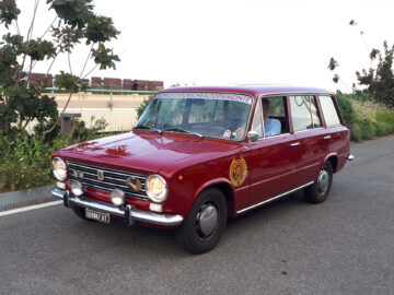 Een rode vintage auto, waarschijnlijk een van de klassieke Fiat-modellen, met een badge op de deur en een bordje op de voorruit met de tekst "CINQUE TORRI CLUB ALTO PIEMONTE", staat geparkeerd op een weg met bomen en een gebouw op de achtergrond. Gespot in deze serene omgeving, het is een prachtig stukje geschiedenis.