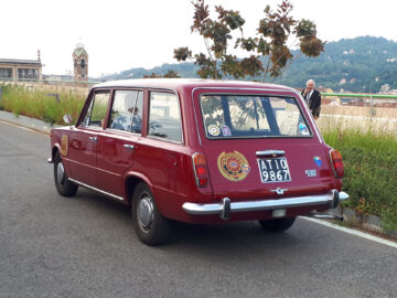 Een kastanjebruine vintage stationwagen, onderdeel van de klassieke Fiat-modellen, staat geparkeerd in een straat met een schilderachtige achtergrond van heuvels en gebouwen. Op het kenteken staat "ATO 9867" en op de achterruit zitten diverse stickers.