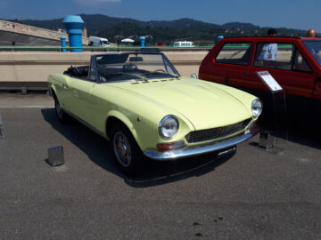 Een vintage lichtgele cabriolet, een van de klassieke Fiat-modellen, wordt op een zonnige dag buiten tentoongesteld met een andere auto en een paar mensen op de achtergrond – gespot tijdens een evenement.