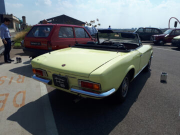 Een lichtgele Fiat cabriolet, die doet denken aan klassieke modellen, staat overdag geparkeerd naast een rode hatchback met het dak open. Er worden drie mannen gezien bij de auto's en in de verte is een brug zichtbaar.