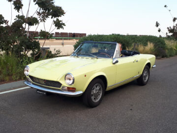 Een gele klassieke cabriolet, die doet denken aan klassieke Fiat-modellen, staat geparkeerd op een weg met bomen en struiken op de achtergrond.