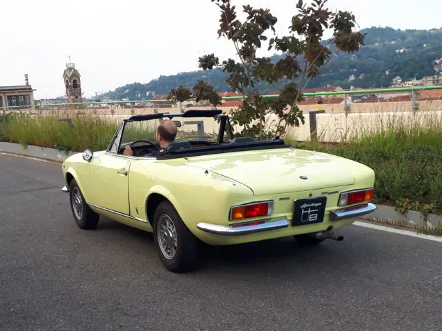 Een persoon rijdt in een lichtgele cabriolet op een dakweg met schilderachtige heuvels en gebouwen op de achtergrond, die doet denken aan klassieke Fiat-modellen.