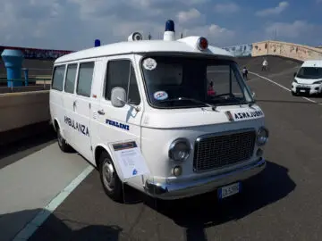 Een vintage witte ambulance met het label "AMBULANZA" geparkeerd op een verhard terrein onder een bewolkte hemel. Voor de klassieke Fiat staat een displaybord.