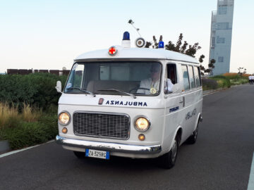 Vintage ambulance met Italiaans kenteken ZA-329RG rijdend op een verharde weg, groen aan de linkerkant en een hoog modern gebouw op de achtergrond. Dit klassieke Fiat-model werd onlangs gespot nabij de binnenstad te midden van het vroege ochtendverkeer.