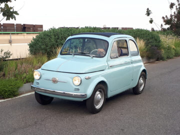 Een lichtblauwe vintage compacte auto, die doet denken aan klassieke Fiat-modellen, rijdt over een verharde weg met groen en een gebouw op de achtergrond. Gespot: elegantie op wielen.